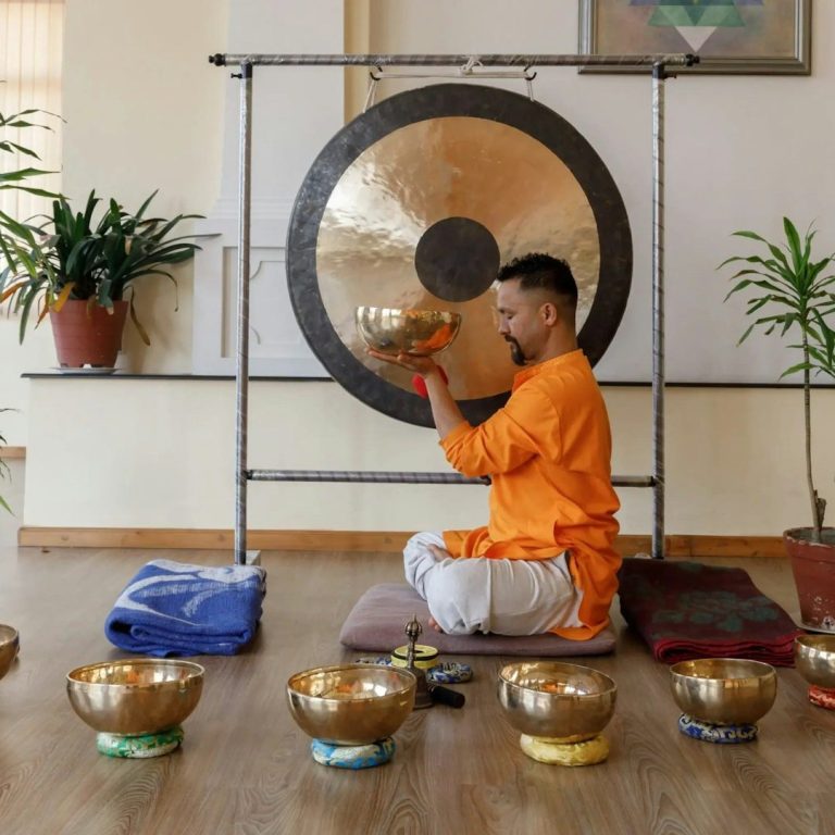Mann in orange Gewand spielt Gong, umgeben von Klangschalen und Pflanzen.